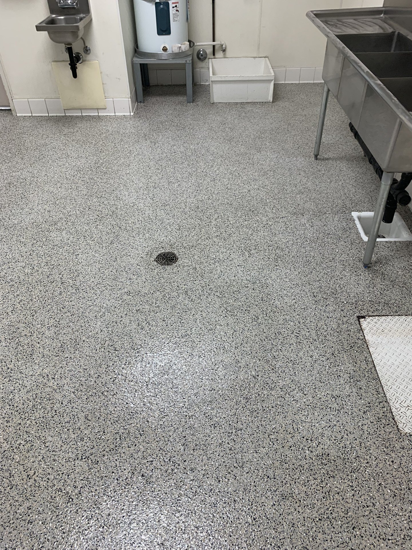 Gray broadcast epoxy floor around a floor drain in a commercial kitchen with a stainless steel three-compartment sink